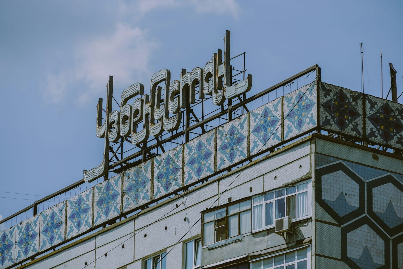 Letrero soviético «Узоры» (motivos decorativos) sobre un edificio con mosaicos, bajo un cielo azul.